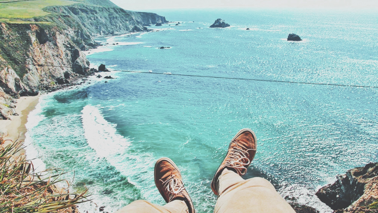 A person sitting on a high cliff overlooks a serene turquoise ocean, with waves gently crashing onto a sandy shore. The view includes rugged coastal cliffs dotted with greenery under a clear sky, as seen from the perspective of the person's brown shoes at the edge of the cliff