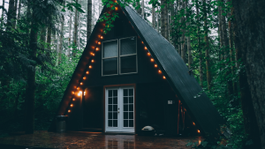 A-frame cabin with string lights nestled in a lush forest setting, showcasing the tranquility of nature-bound architecture on a damp wooden deck